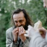 A man happily eats a sandwich outside in a green and natural setting.